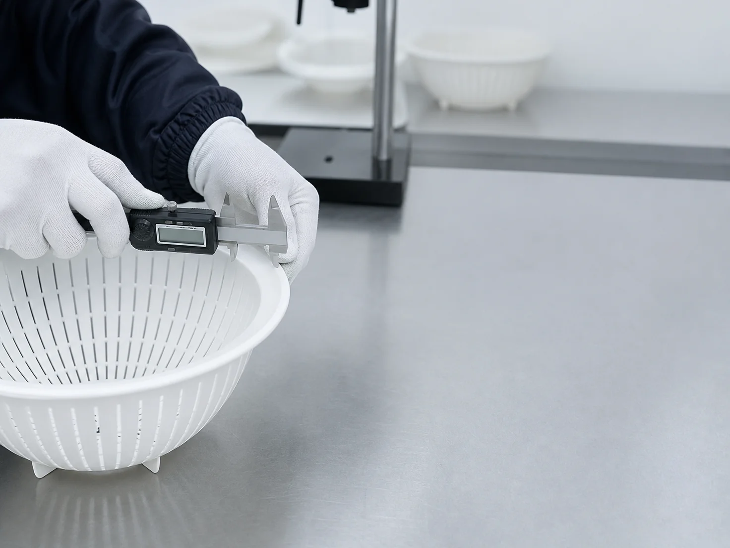 Gloved hands measure a white plastic colander with a digital caliper at an inspection table.